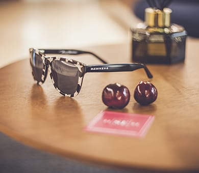 Gafas de sol Hawkers, perfume y cerezas en mesa