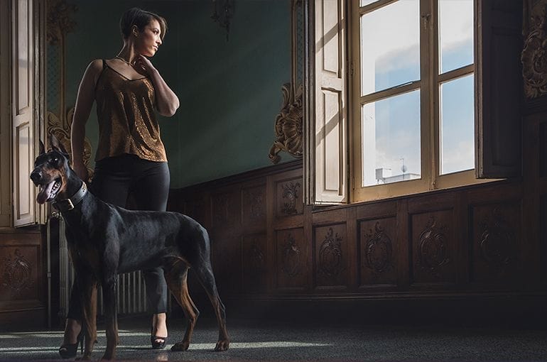 Mujer con vestido dorado y perro negro en habitaci&oacute;n con ventanas.