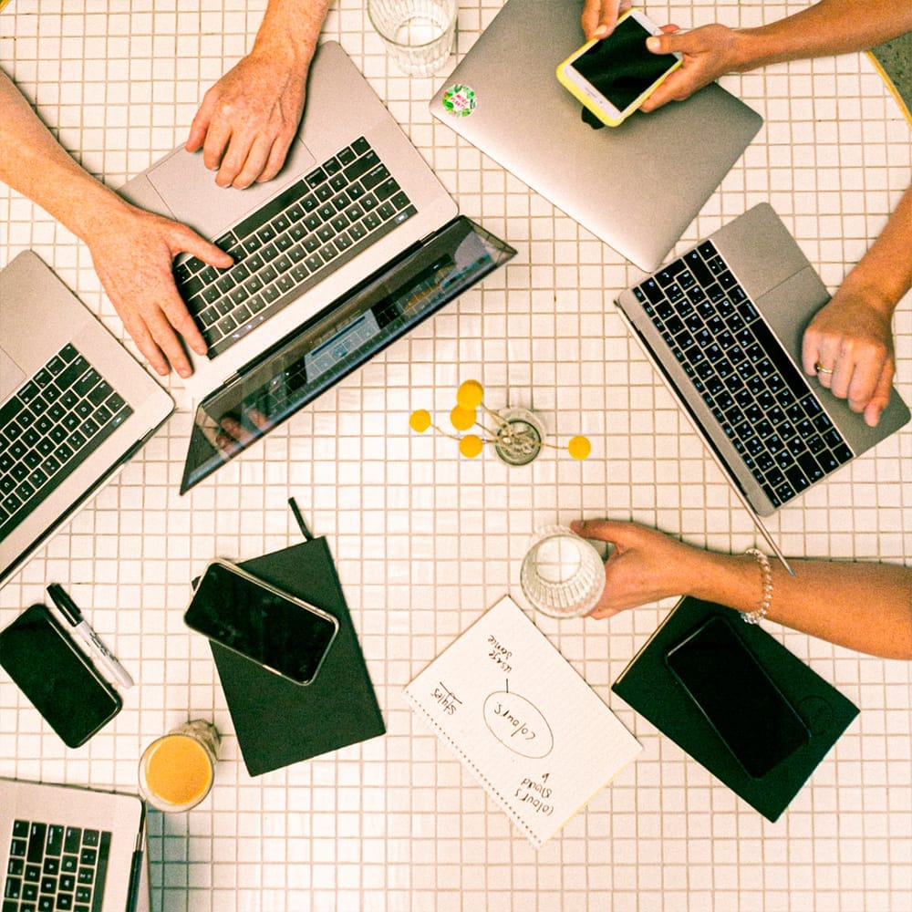 Cuatro personas trabajando en una mesa con ordenadores portátiles y teléfonos.